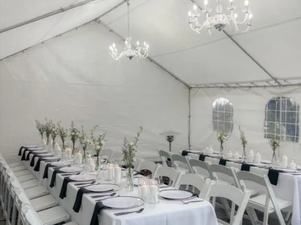 Long banquet tables with white linens and black napkins under chandeliers in a white tent