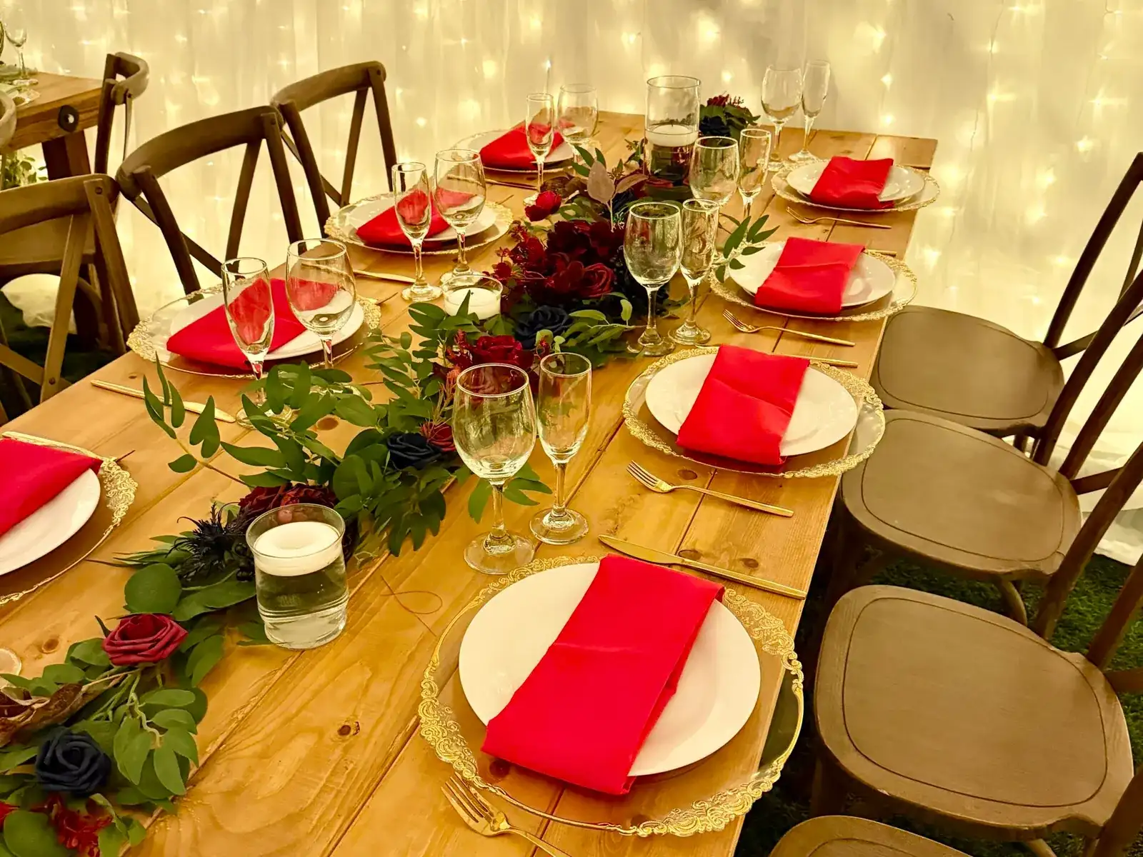 Wooden farm table set with red napkins, gold chargers, and greenery in front of a wall of fairy lights