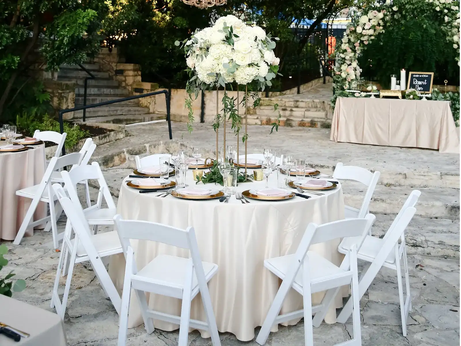Round tables with white folding chairs and a tall white floral centerpiece on a stone patio